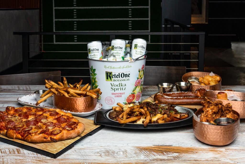 A variety of food items on a table: pizza, fries, wings, and pretzels. There is also a bucket full of Ketel One vodka cans.