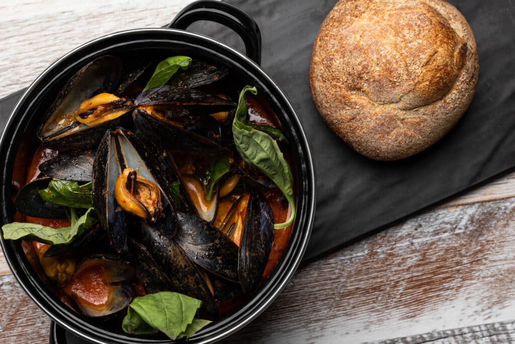 A black saucepan sits on a table. It contains cooked mussels and seasonings. A dinner roll sits next to the saucepan.